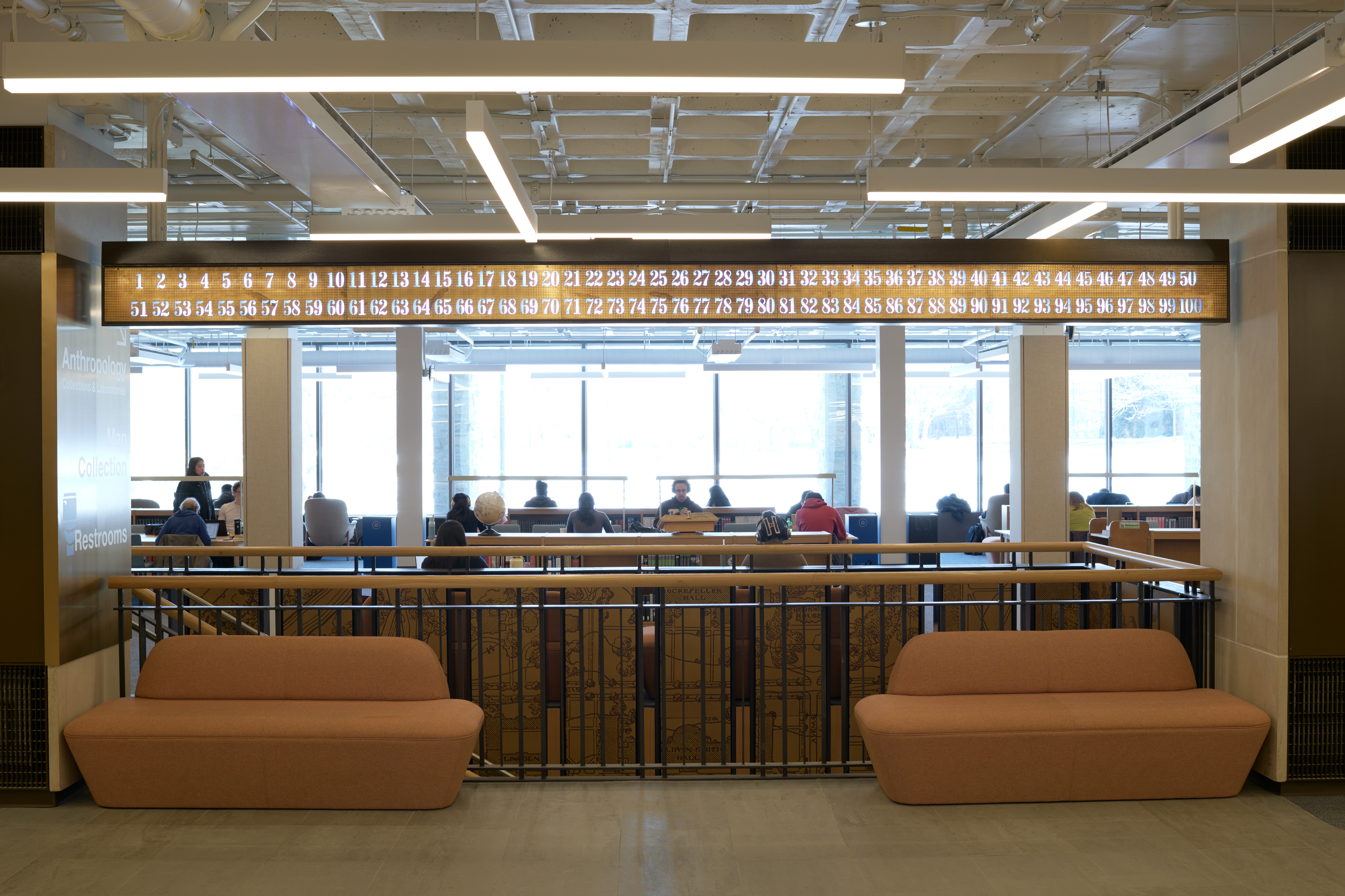 The call board in Olin Library is placed between two concrete pillars, above a stair case. Tan couches are in front of the staircase and students can be seen studying in the background. 