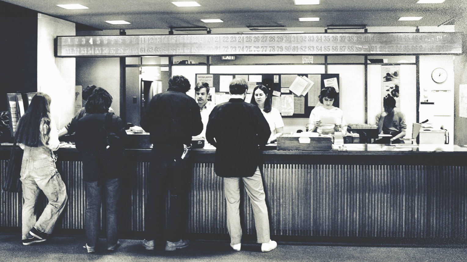 Staff help students at the library circulation desk. The call board is above the desk with a few numbers illuminated. 
